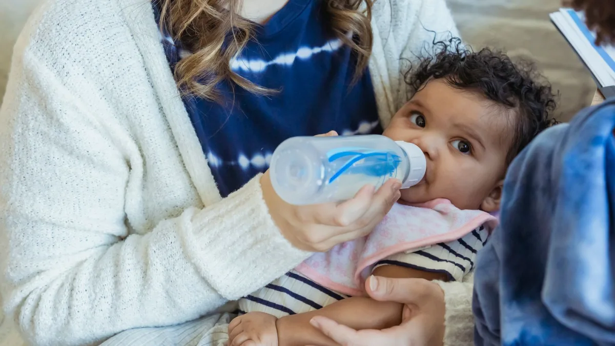 A parent bottle-feeding a baby on a couch in a warm, softly lit home