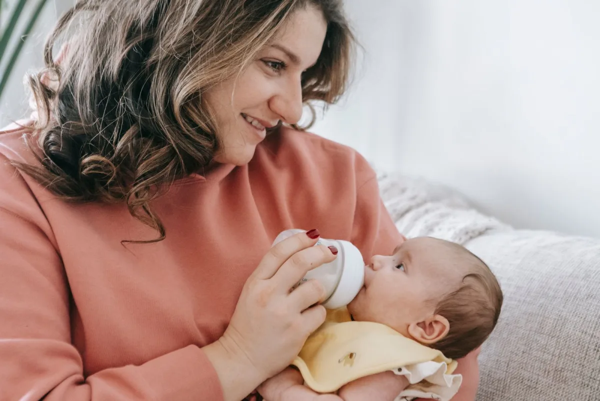 A smiling mother feeding her newborn a prepared bottle of formula in soft warm light